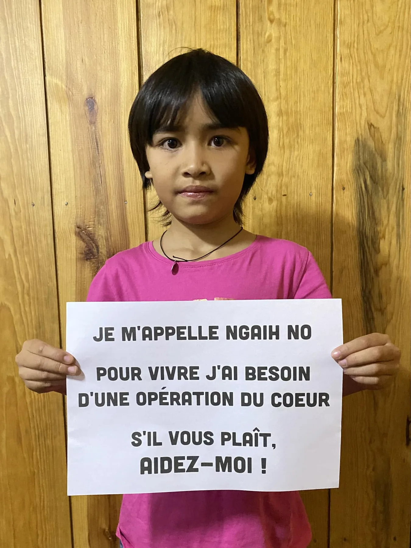 Ngaih No tenant une pancarte : « Je m'appelle Ngaih No. Pour vivre j'ai besoin d'une opération du cœur. S'il vous plaît, aidez-moi ! »