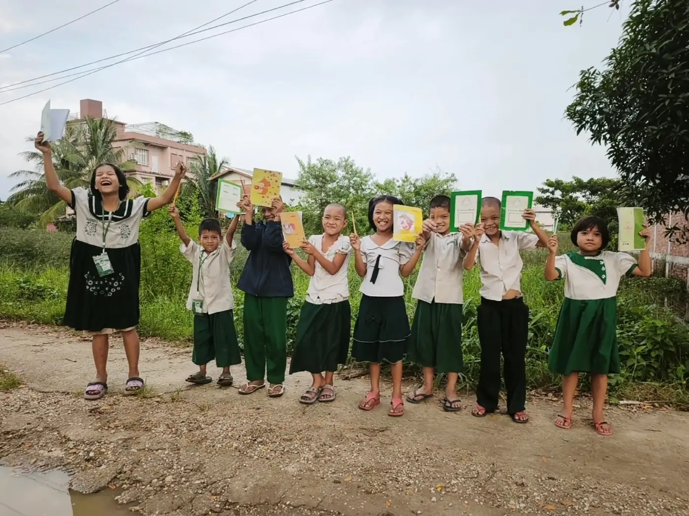 Nine children from the Drop-In Center in a line on a dirt track, holding up their exercise books and pencils